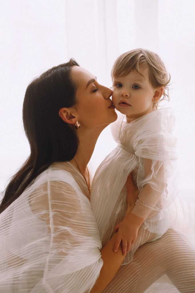 Happy mother in white dress sitting near window, holding her bab